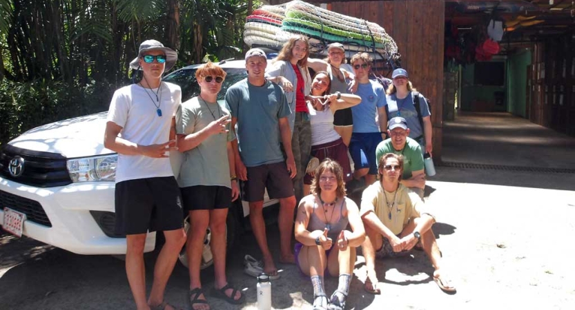 A group of students stand beside a car with surfboards on the roof. 